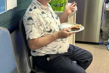 Thumbnail for Man with glasses in patterned shirt sitting and eating a burger at an indoor gathering with drinks and red cups nearby.