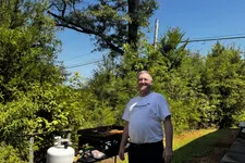 Thumbnail for Man standing near a grill with propane tank in a sunny backyard surrounded by green trees and blue sky.