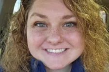 Thumbnail for Smiling woman with curly hair wearing a blue zip-up jacket with a logo, sitting inside a car.