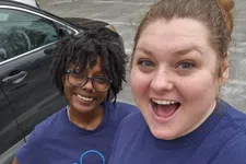 Thumbnail for Two smiling women wearing matching blue Abiomed T-shirts, one giving a thumbs up, outdoors in a parking lot.
