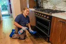 Thumbnail for Woman cleaning oven door inside modern kitchen using spray cleaner and cloth, smiling while kneeling on wooden floor