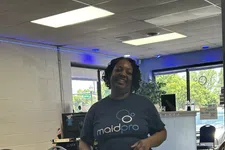Thumbnail for Smiling woman wearing a MaidPro t-shirt standing in a room with tables, chairs, and office supplies.