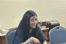 Thumbnail for Young woman with long dark hair sitting at a table with a bowl of sliced cucumbers, red cup, and soda bottle.