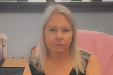 Thumbnail for Woman with blonde hair wearing a black sleeveless shirt with colorful puzzle flower design sitting at a wooden desk.