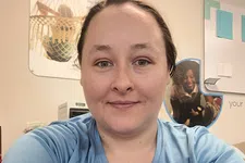 Thumbnail for Smiling woman in light blue shirt takes a selfie in an office with posters and papers on the wall behind her.