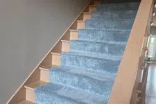 Thumbnail for Indoor wooden staircase covered with soft blue-gray carpet runner and light brown handrail.