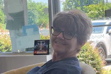 Thumbnail for Smiling woman wearing glasses sitting in a chair near a window with outdoor greenery and a standing cutout figure visible.