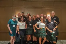 Thumbnail for Group of eleven people smiling indoors, most wearing green tartan kilts and casual tops, posing in front of wood panel wall.