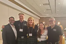 Thumbnail for Five people smiling indoors wearing matching green tartan kilts and name badges at an event.