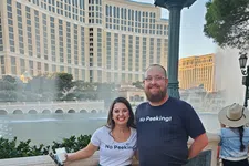 Thumbnail for Smiling couple wearing matching green plaid kilts and No Peeking shirts by a fountain with hotel buildings in the background