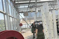 Thumbnail for Two people in kilts inside a greenhouse with vertical hydroponic towers and red hose reel.