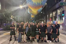 Thumbnail for Group of people wearing kilts standing on a city street at night under colorful hanging art installation.