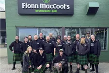 Thumbnail for Group of people wearing green plaid kilts posing outside Fionn MacCool's restaurant and pub on a city sidewalk.