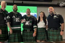 Thumbnail for Group of six people wearing green plaid kilts and black shirts, smiling in front of a green screen backdrop.