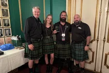 Thumbnail for Four people wearing green plaid kilts posing indoors, smiling and holding an award, standing in ornate room.