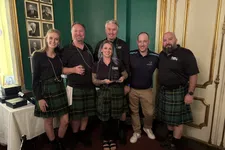Thumbnail for Group of six people, five wearing green tartan kilts, posing indoors with award in decorated room.