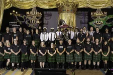 Thumbnail for Group photo of men and women wearing green plaid kilts and black shirts posing on stage under gold curtains and chandeliers.