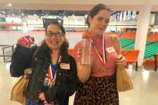 Thumbnail for Two women wearing medals smile and hold trophies inside an indoor sports arena with orange and green seats.