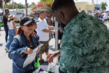 Thumbnail for Woman in denim jacket and cap engaging with man in green shirt at outdoor event booth on sunny day