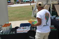 Thumbnail for Person browsing an outdoor vendor table with promotional items and brochures on a sunny day.