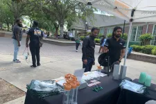 Thumbnail for Outdoor campus event with information booth offering promotional items and two men staffing the table under a canopy.