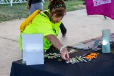 Thumbnail for Woman in neon yellow shirt arranging promotional items on a black table at an outdoor event.