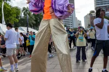 Thumbnail for Man on stilts wearing shiny purple jacket, orange shirt, gold pants, and afro wig at outdoor city event.