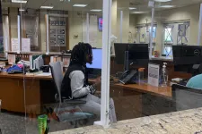 Thumbnail for Office reception desk with pens, a water bottle, acrylic divider, and a person working on a computer.