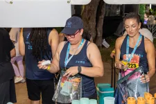 Thumbnail for Two women wearing Zuma shirts distributing cups and water bottles at an outdoor event booth.