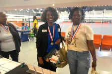 Thumbnail for Two women wearing medals holding gift bags at an indoor event with orange seating in the background