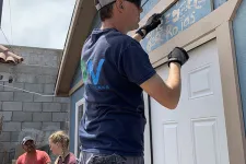 Thumbnail for Man wearing cap and gloves painting a wooden sign above a white door on a blue house with two people conversing nearby.