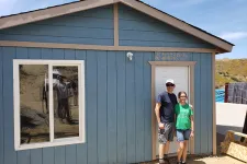 Thumbnail for Man and girl standing in front of a small blue wooden house with a white door and window on a sunny day.