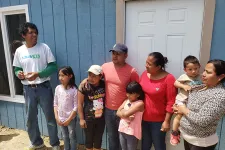 Thumbnail for Group of seven people, including adults and children, standing outside a blue house near a white door on a sunny day