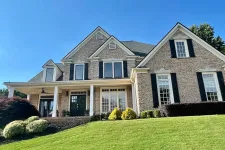 Thumbnail for Large brick suburban house with black shutters and a manicured green lawn under a clear blue sky.