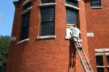 Thumbnail for Man on ladder painting or cleaning a large curved red brick building with tall windows under a clear blue sky