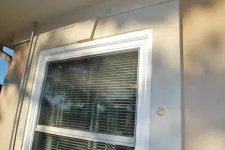 Thumbnail for White-framed window with horizontal blinds on beige stucco wall reflecting trees and sky outdoors.