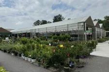Thumbnail for Outdoor plant nursery with various potted shrubs and flowers displayed around a large greenhouse under a partly cloudy sky.