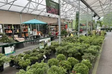 Thumbnail for Indoor garden center with rows of evergreen shrubs, potted plants, patio furniture, and gardening supplies under a large greenhouse roof.