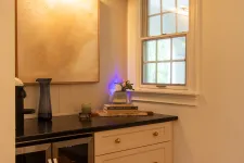 Thumbnail for Cozy kitchen corner with black countertop, wine fridge, cream cabinets, decorative art, and window letting natural light in.