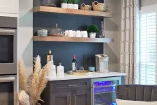 Thumbnail for Modern kitchen corner with double stainless steel oven, dark cabinetry, open shelves, and cozy gray armchair near window shutters.