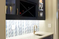 Thumbnail for Modern wet bar with navy blue cabinets, wine rack, white countertop, brass faucet, and wave-patterned backsplash.