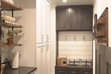 Thumbnail for Modern laundry room with dark wood storage bench, white cabinets, herringbone tile floor, and rustic ceiling beam