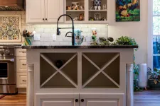 Thumbnail for Modern kitchen island with farmhouse sink, white cabinetry, hardwood floors, and decorative shelves with plants and art.