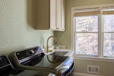 Thumbnail for Bright laundry room with modern black washer and dryer, white cabinetry, patterned wallpaper, and large window.
