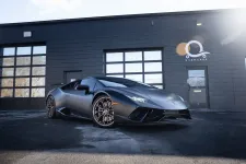 Thumbnail for Matte black Lamborghini sports car parked in front of a black garage with glass windows on a sunny day.