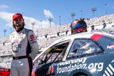 Thumbnail for Race car driver in white suit and red cap stands next to black and red number 60 stock car on racetrack