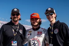Thumbnail for Three men smiling at a race track, with one in a racing suit and two in black hoodies under clear blue sky.
