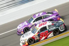Thumbnail for Two stock cars, number 90 in black and white and number 77 in purple, race side by side on a track at high speed.