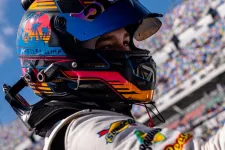 Thumbnail for Race car driver wearing a colorful helmet and white suit at a motorsport event with grandstands in the background