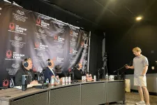 Thumbnail for Three men with headsets sit at a panel table recording a podcast while a young man stands holding a microphone boom.